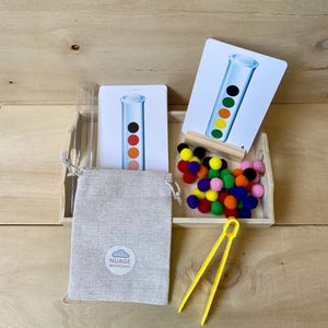 May include: A Montessori learning set in a wooden tray. It features cards with test tube illustrations, colourful pom-poms, a small bag with the "Nuage Montessori" logo, a clear tube, and yellow tongs. The cards show coloured dots.
