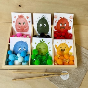 Peut inclure: Un plateau de tri en bois avec six compartiments, chacun contenant une carte illustrée d'un monstre coloré et des pompons assortis en rose, noir, rouge, bleu, vert et orange. Une paire de pinces en bambou et un petit sac en toile sont inclus.