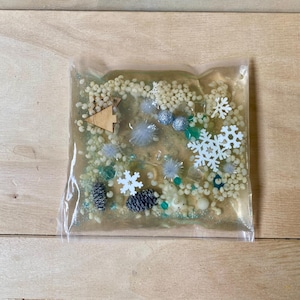Op de afbeelding: Een doorzichtige, verzegelde sensorische zak gevuld met kleine witte kralen, sneeuwvlokken en decoratieve elementen. Bevat een houten boom, zilveren en blauwe ornamenten en kleine dennenappels. De zak ligt op een houten oppervlak.