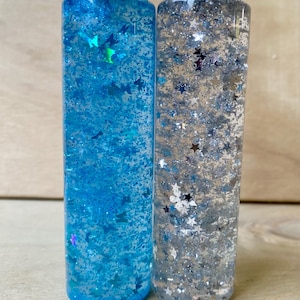 Op de afbeelding: Twee heldere sensorische flessen gevuld met glitter en vormen. Eén fles heeft blauwe vloeistof met blauwe en iriserende vlindervormen. De andere heeft zilveren vloeistof met zilveren stervormen. Beide hebben zilveren doppen.