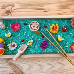 Può includere: Un vassoio di legno riempito di riso verde, decorato con fiori colorati, coccinelle, api e pompon. Include un cucchiaio di legno e pinzette di bambù per il gioco sensoriale. Un'attività divertente e coinvolgente per i bambini.