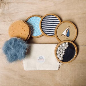 Op de afbeelding: Een verzameling nautische onderzetters met houten randen, met ontwerpen zoals een zeilboot, blauw golfpatroon en marineblauwe strepen. Inclusief een blauw pluizig item, een spons en een kleine canvas tas.