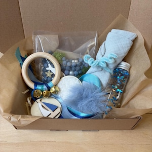 Peut inclure: Un kit sensoriel dans une boîte en carton contenant un anneau en bois avec des grelots dorés, une bouteille transparente remplie de liquide à paillettes bleues, un rouleau de tissu bleu clair, une découpe de voilier en bois et un pompon bleu.