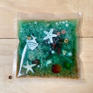 Op de afbeelding: Een doorzichtige plastic zak gevuld met een groene gel, zand en diverse decoratieve elementen. De zak bevat witte zeesterren, een witte vis, parels en kleine schelpen. Het algemene thema is geïnspireerd op de oceaan.