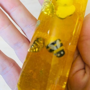 May include: A clear, rectangular resin piece filled with yellow liquid and small bee-shaped embellishments. The bees are yellow and black. The resin piece is held in a hand.