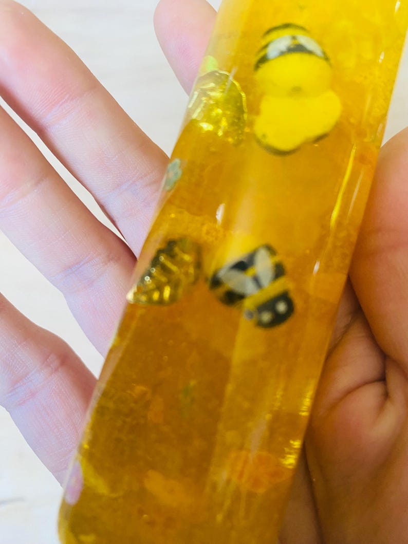 May include: A clear, rectangular resin piece filled with yellow liquid and small bee-shaped embellishments. The bees are yellow and black. The resin piece is held in a hand.