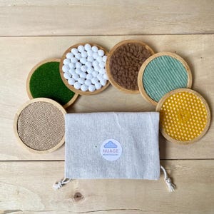 Op de afbeelding: Een set van zeven ronde onderzetters met verschillende texturen, waaronder groen vilt, witte pompons, bruin imitatiebont en gele honingraatstof. Een kleine grijze stoffen zak met een label "Nuage Montessori" is ook inbegrepen.