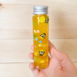 May include: A clear, cylindrical bottle with a silver cap filled with yellow liquid and decorative elements. The bottle features small bee and flower-shaped embellishments. The bottle is held in a hand against a light wood background.