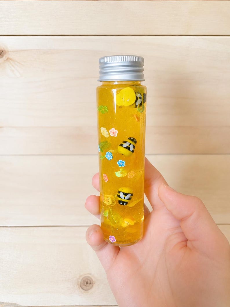 May include: A clear, cylindrical bottle with a silver cap filled with yellow liquid and decorative elements. The bottle features small bee and flower-shaped embellishments. The bottle is held in a hand against a light wood background.