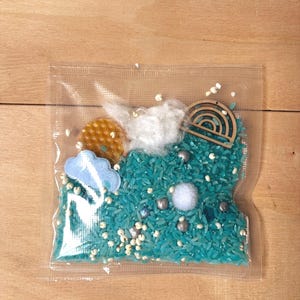 Op de afbeelding: Een doorzichtige plastic zak gevuld met in talingkleur geverfde rijst, witte watten, een kleine houten regenbooguitsparing, een gele schijf met honingraatstructuur, een lichtblauwe viltwolk, een witte pompon en diverse kleine zilveren en witte kralen.