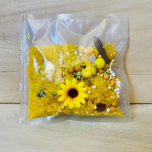 May include: A clear plastic bag filled with yellow rice, a yellow sunflower, and small bee-shaped toys. The bag also contains small yellow and white beads, and a wooden stick. The contents are designed for sensory play.