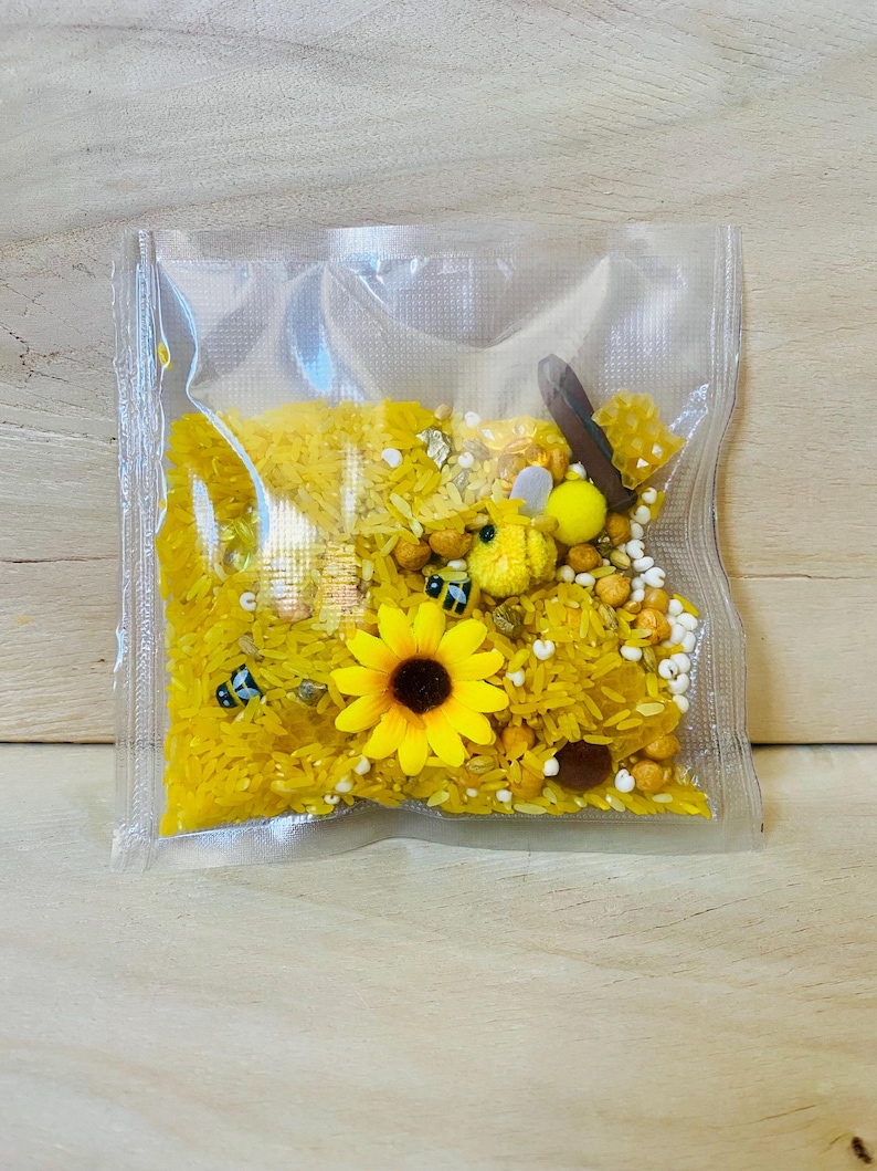 May include: A clear plastic bag filled with yellow rice, a yellow sunflower, and small bee-shaped toys. The bag also contains small yellow and white beads, and a wooden stick. The contents are designed for sensory play.