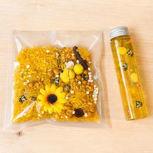 May include: A sensory kit with yellow rice, sunflower, and bee-themed elements in a clear bag. A clear bottle with yellow liquid and bee and flower decals is next to the bag. The items are on a light wood surface.