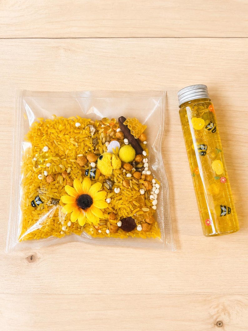 May include: A sensory kit with yellow rice, sunflower, and bee-themed elements in a clear bag. A clear bottle with yellow liquid and bee and flower decals is next to the bag. The items are on a light wood surface.