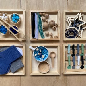 May include: A selection of wooden trays containing craft materials. Features small bowls with pom-poms, wooden beads, pipe cleaners, socks, a bowl with blue gems, wooden spoons, star-shaped wooden cutouts, and pegs.