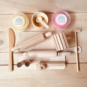 Peut inclure: Un ensemble de jeu en bois avec un rouleau à pâtisserie, une planche à découper et divers outils. Comprend deux pots de pâte à modeler, un jaune et un rose, avec le texte "NUAGE MONTESSORI". L'ensemble est disposé sur une surface en bois clair.