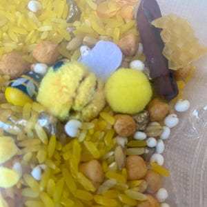 May include: A sensory bin filled with yellow dyed rice, small beige and brown beans, white beads, yellow pom-poms, and a small felt heart. There is also a small, dark brown object and a yellow honeycomb-shaped object.