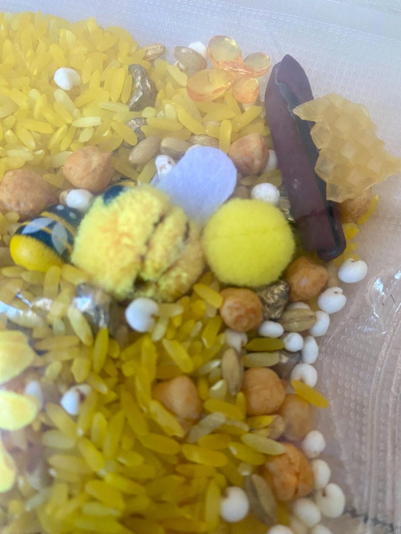 May include: A sensory bin filled with yellow dyed rice, small beige and brown beans, white beads, yellow pom-poms, and a small felt heart. There is also a small, dark brown object and a yellow honeycomb-shaped object.