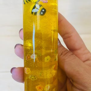 May include: A clear, cylindrical bottle filled with yellow liquid and glitter. The bottle contains small, decorative bees and flower shapes. The bottle is held by a person.