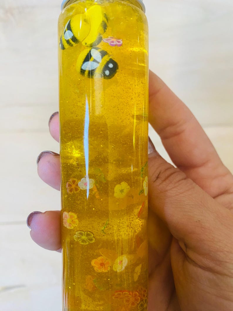May include: A clear, cylindrical bottle filled with yellow liquid and glitter. The bottle contains small, decorative bees and flower shapes. The bottle is held by a person.