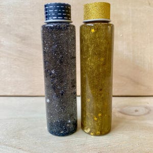 Op de afbeelding: Twee heldere, cilindrische sensorische flessen gevuld met glitter. Eén fles bevat zwarte glitter met kleine stervormige confetti, de andere heeft gouden glitter met stervormige confetti. De flessen hebben decoratieve doppen.
