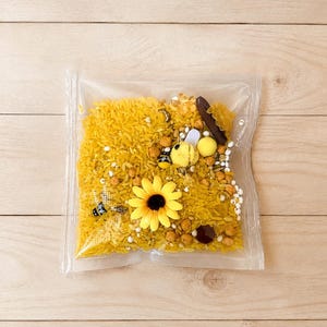 May include: A clear plastic bag filled with yellow dyed rice, small beads, and decorative elements. A yellow sunflower and a small bee figurine are visible. The bag is on a light wood surface.