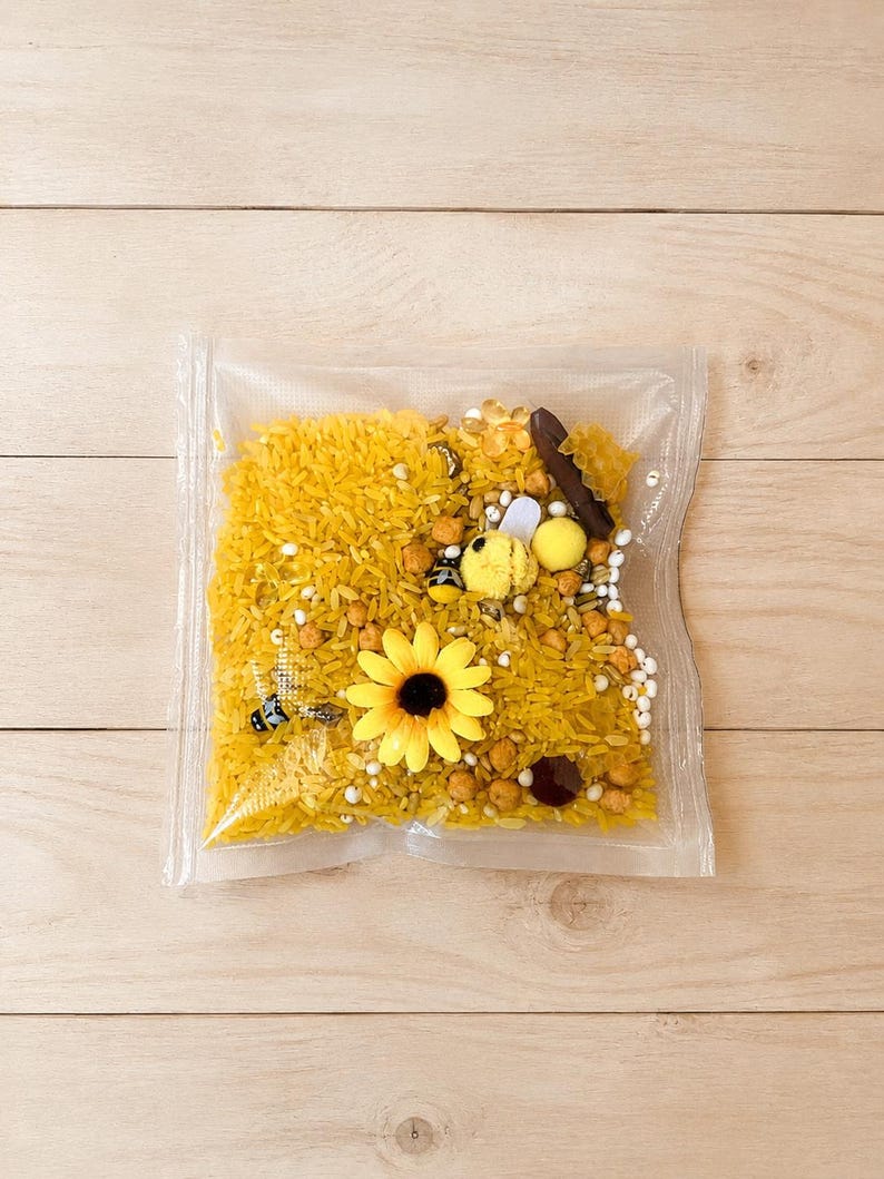 May include: A clear plastic bag filled with yellow dyed rice, small beads, and decorative elements. A yellow sunflower and a small bee figurine are visible. The bag is on a light wood surface.