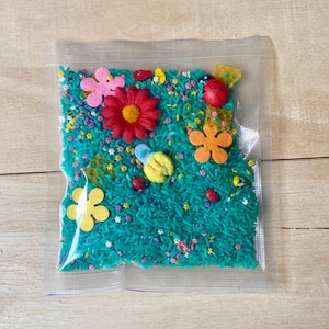 Op de afbeelding: Een sensorische activiteitenzak gevuld met turquoise gekleurde rijst, versierd met vilten bloemen, bijen en lieveheersbeestjes. De zak bevat een verscheidenheid aan kleurrijke vormen en texturen, perfect voor tactiele verkenning en spel.