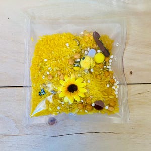 May include: A clear plastic bag filled with yellow dyed rice, small wooden pieces, and decorative elements. The bag contains a yellow sunflower, small bee figurines, and white beads. The contents are arranged on a light wooden surface.