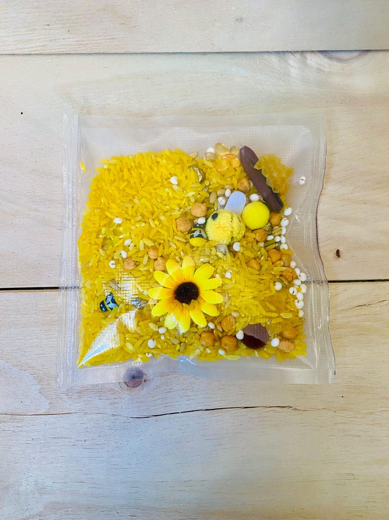 May include: A clear plastic bag filled with yellow dyed rice, small wooden pieces, and decorative elements. The bag contains a yellow sunflower, small bee figurines, and white beads. The contents are arranged on a light wooden surface.