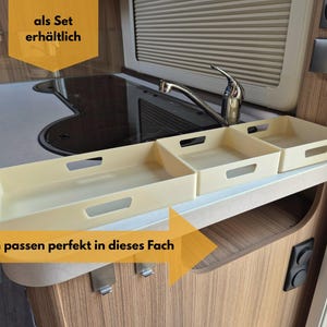 May include: Three off-white rectangular storage trays with handle cutouts are arranged on a shelf. The trays are in a kitchen setting, near a sink and countertop. Text in the image says "als Set erhältlich" and "Boxen passen perfekt in dieses Fach."