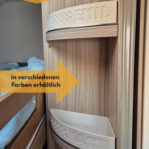 May include: Two white, curved shelves with the names "EMIL" and "MATTI" embossed on them. The shelves are mounted on a wooden panel. Text in the image reads "personalizierbar" and "in verschiedenen Farben erhältlich".