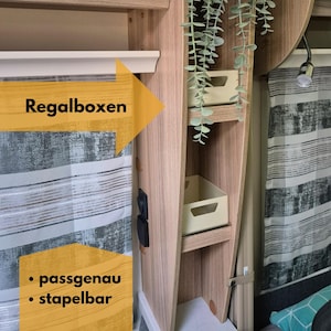 May include: Wooden shelving unit with two beige storage boxes. The unit is built into a wall with a patterned curtain. Text on a yellow arrow reads "Regalboxen", with the words "passgenau" and "stapelbar" below.