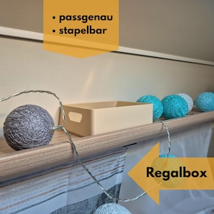 May include: A beige rectangular storage box with a handle is on a wooden shelf. Decorative string lights with gray and blue spheres are strung along the shelf. The text "Regalbox" and "passgenau, stapelbar" are in the image.