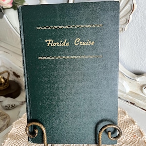 May include: A dark green book titled "Florida Cruise" with gold lettering and decorative lines. The book is displayed on a decorative, ornate, gold-colored metal stand, set against a neutral background.