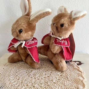 May include: Two brown plush rabbits with long ears and black eyes. Each rabbit wears a red cape with white scalloped trim and white ribbon ties. The left rabbit has the name Cotton-tail embroidered in white, and the right rabbit has Topsy.