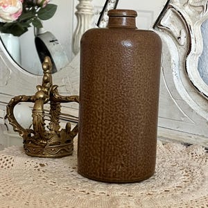 May include: A brown, textured ceramic bottle with a small neck and a rounded body. A gold-colored crown sits to the left of the bottle. The bottle is on a lace doily.