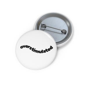 May include: A round white pin-back button featuring the word overstimulated written in a black, textured, sans-serif font across the center. The back of the button is metallic silver and includes a horizontal pin mechanism for attachment.