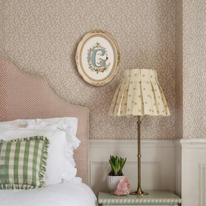 Può includere: Una scena di camera da letto con una testiera rosa a fantasia, biancheria da letto bianca e un cuscino a quadretti verdi. Un'opera d'arte ovale incorniciata con la lettera "G" è appesa sopra un comodino con una lampada e una piccola pianta in vaso. La lampada ha un paralume plissettato.