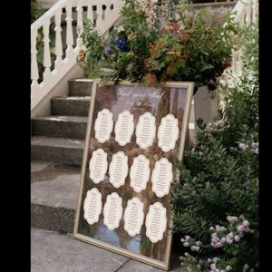 May include: A framed seating chart with the text "Find your Seat" displayed at an outdoor wedding. The chart features several individual name cards with decorative borders. The background shows a landscape scene with trees and a staircase.