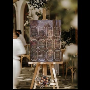 May include: A wooden easel displays a seating chart with the text "Find your seat". The chart features a dark background with ornate frames, each containing guest names. A floral arrangement rests at the base of the easel, adding a decorative touch.