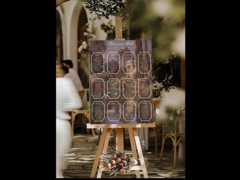 May include: A wooden easel displays a seating chart with the text "Find your seat". The chart features a dark background with ornate frames, each containing guest names. A floral arrangement rests at the base of the easel, adding a decorative touch.