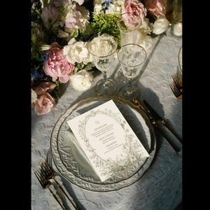 May include: A formal table setting with a menu card, plates, and silverware. The menu card is white with a floral border. The plates are light blue with a gold rim. Two wine glasses are visible, along with a fork and knife. A floral arrangement is in the background.