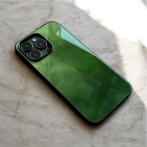 Peut inclure: Coque de smartphone verte avec une finition brillante et un bord noir. Le boîtier présente un module caméra avec trois lentilles. L'étui de téléphone est posé sur une surface claire, avec des ombres de la lumière du soleil.