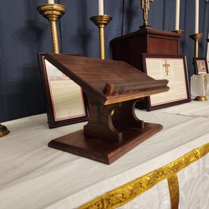 May include: A dark wooden lectern with a sloping top and a decorative base. The lectern is on a white tablecloth with gold trim. Gold candle holders and framed pictures are in the background. The lectern is likely for religious use.