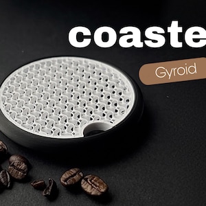 May include: A black and white coaster with a unique patterned design, labeled "coaster" in white text. The coaster is next to a pile of coffee beans. The word "Gyroid" is in a brown rectangle.