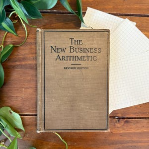 May include: A vintage hardcover book titled The New Business Arithmetic Revised Edition with a tan fabric cover and black text, featuring a thin black border frame on the front cover, positioned alongside white grid paper and green foliage.
