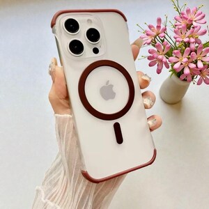 Può includere: Custodia trasparente per iPhone con bordo marrone e anello MagSafe circolare marrone. La custodia ha un interno bianco e presenta il logo Apple. La custodia è mostrata con un piccolo vaso di fiori rosa.