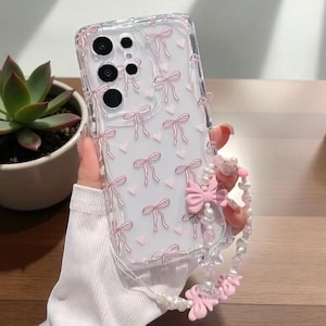 Könnte beinhalten: Transparente Handyhülle mit einem Muster aus rosa Schleifen und Herzen. Sie hat eine Handschlaufe mit Perlen und rosa Schleifen. Die Hülle liegt auf einer Holzoberfläche, im Hintergrund ist eine Sukkulente zu sehen.