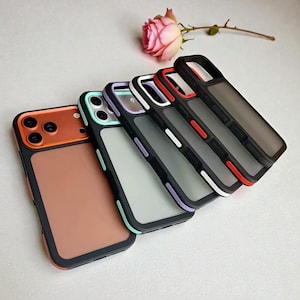 Puede incluir: Una colección de cinco fundas para teléfonos móviles en varios colores. Las fundas tienen un borde negro y una parte trasera esmerilada. Los colores incluyen naranja, verde menta, morado, blanco y rojo. Una rosa rosa en el fondo.